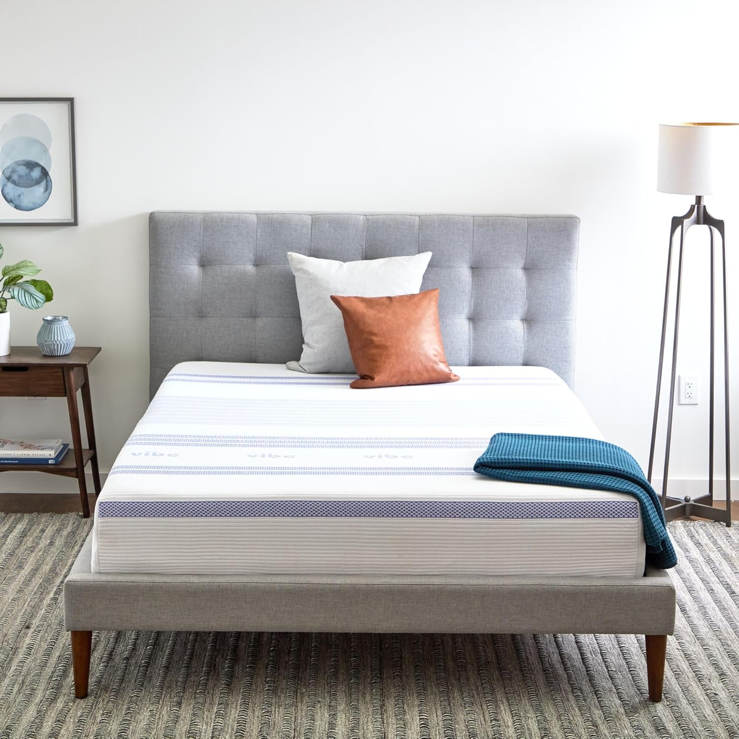 Front view of a mattress on a gray upholstered bed with pillows, a teal throw, and bedside lamp in a bright room.
