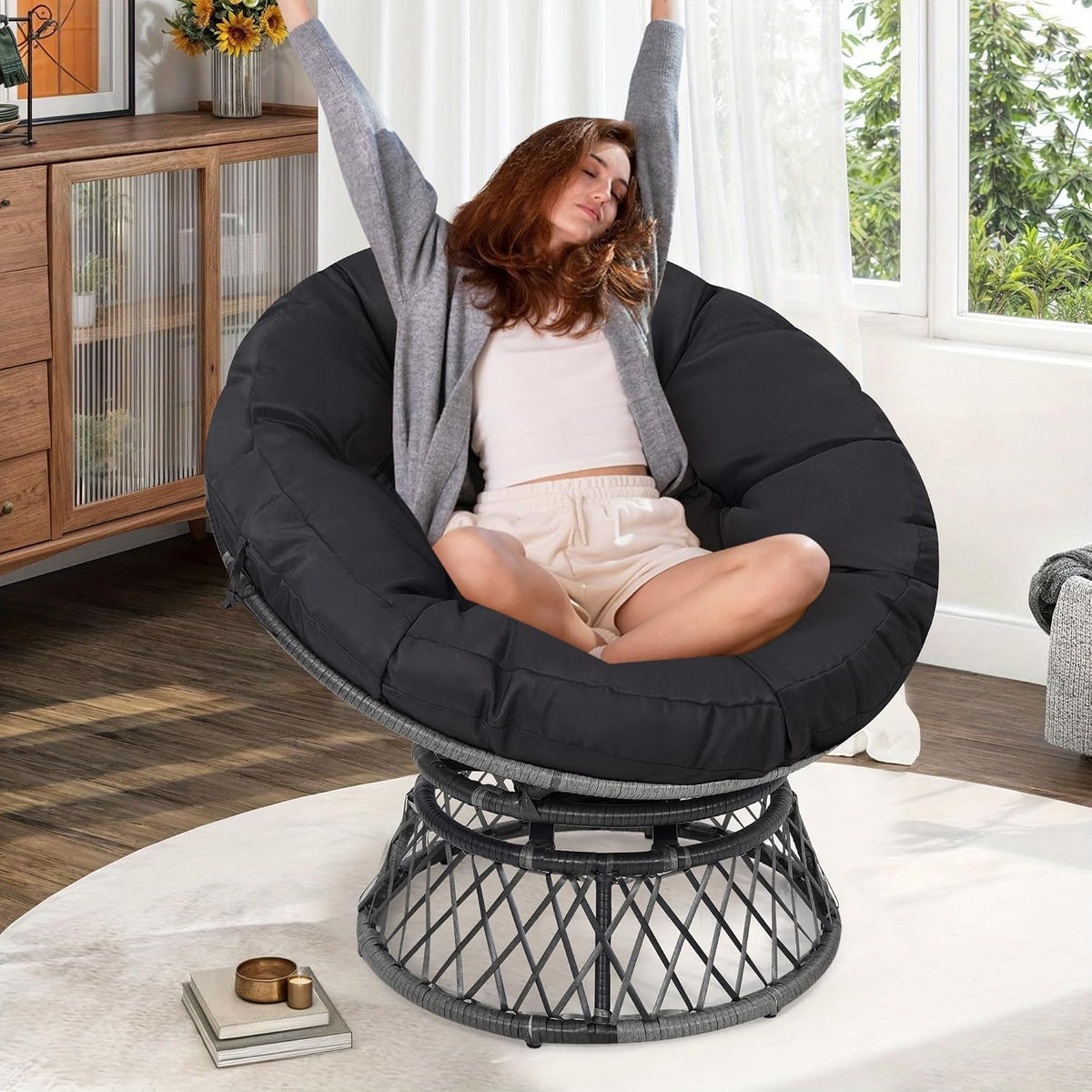 Woman lounging in a round papasan chair with thick black cushion and grey woven base in a living room.
