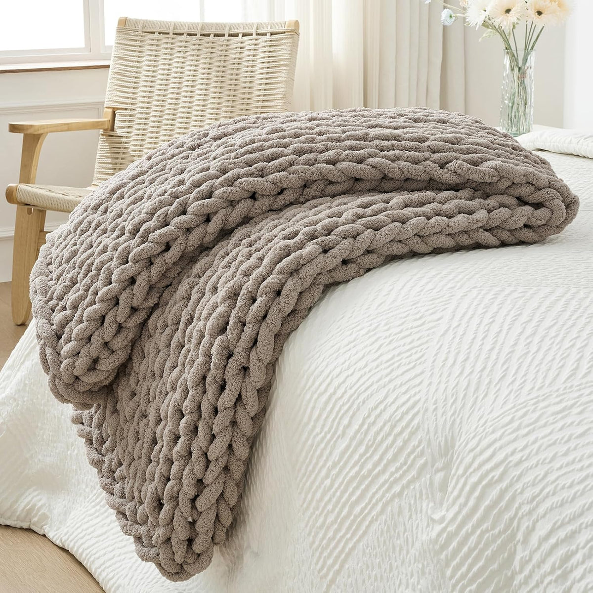 Taupe chunky knit throw loosely folded on a white textured duvet with a woven chair and flowers in background.