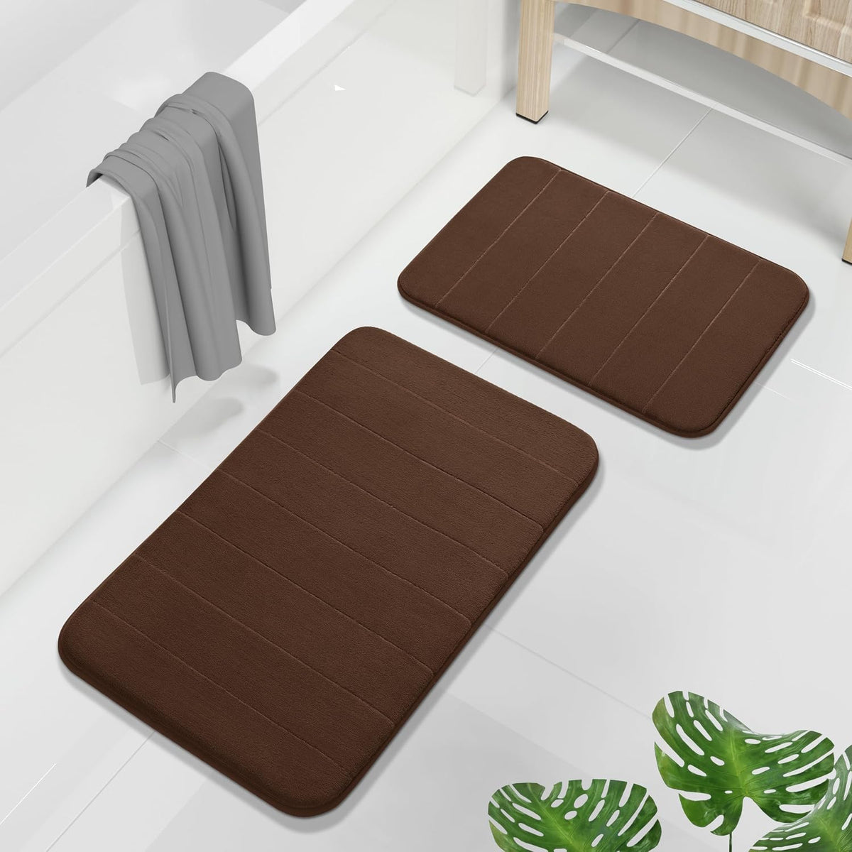 Two brown rectangular bathroom mats on glossy white tile beside a bathtub and wooden vanity.