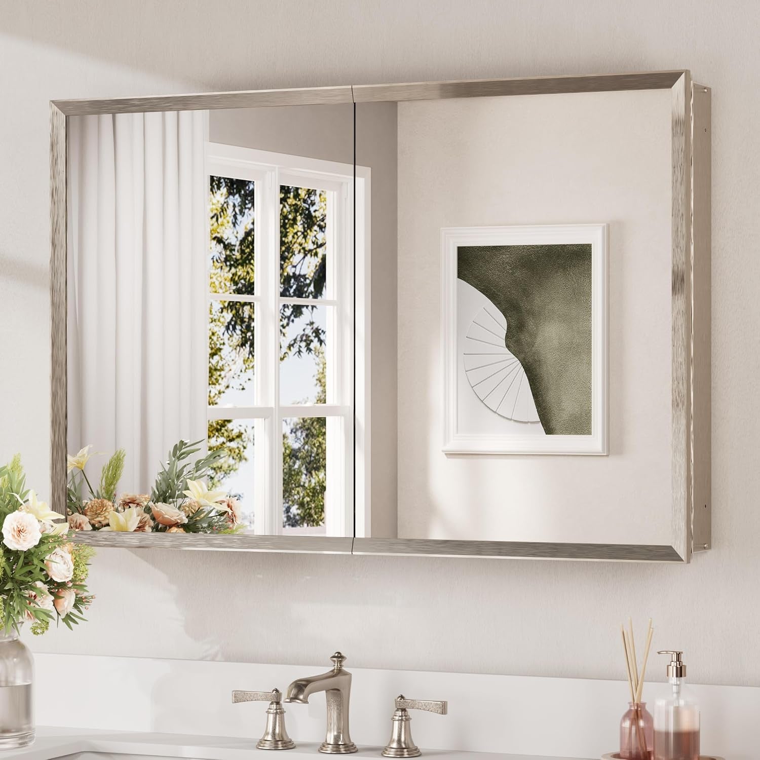 Wall-mounted mirrored medicine cabinet with a brushed silver frame reflecting a window and floral arrangement