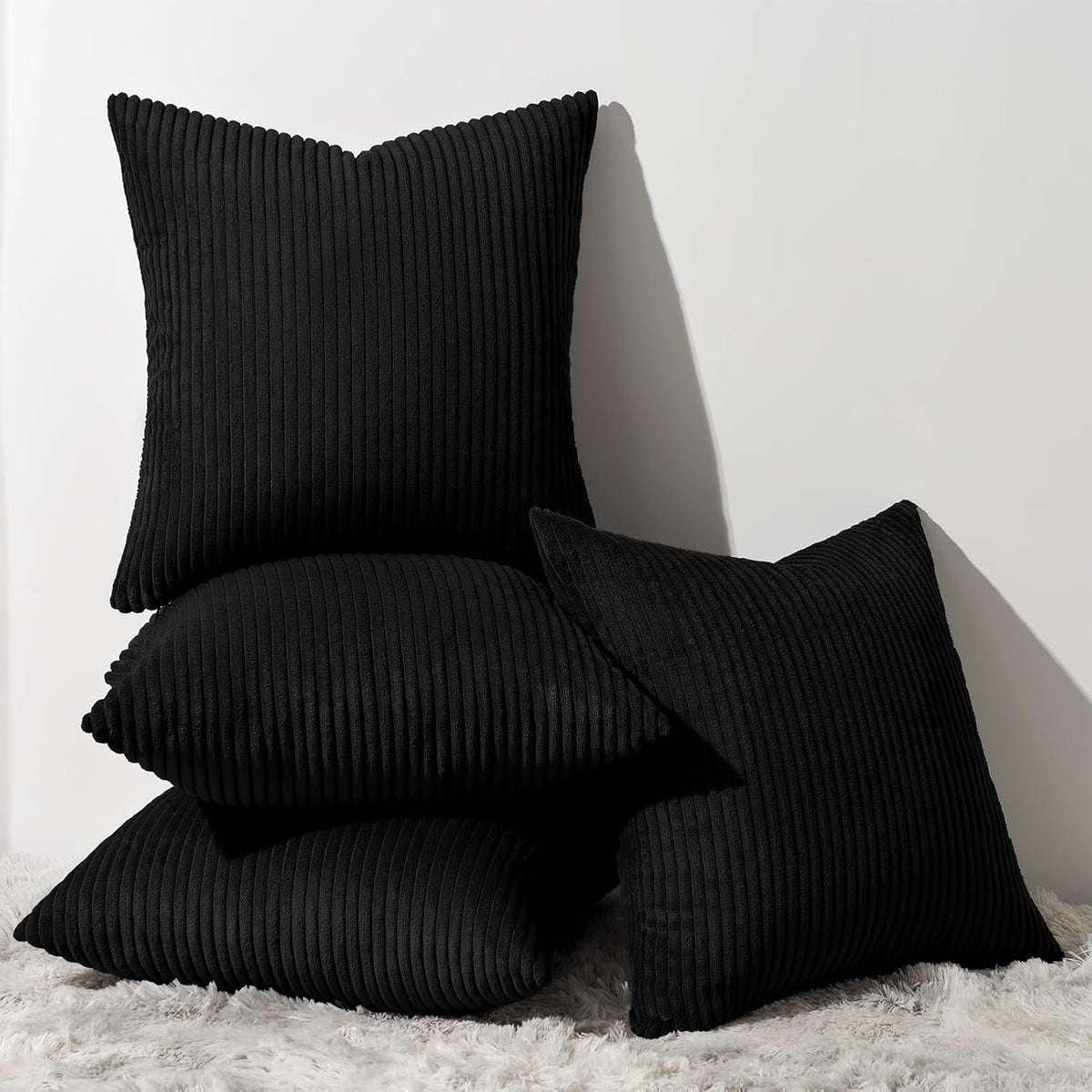 Stack of four black ribbed corduroy square pillows against a white wall on a shag rug.