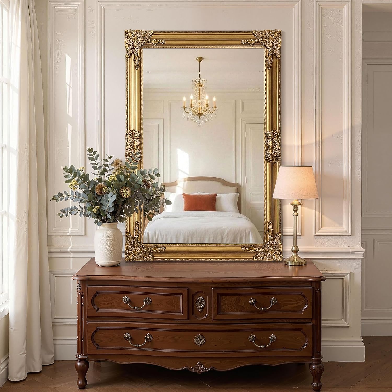 Large ornate gold mirror placed on a wooden dresser reflecting a bed and chandelier, with a vase and lamp at sides.