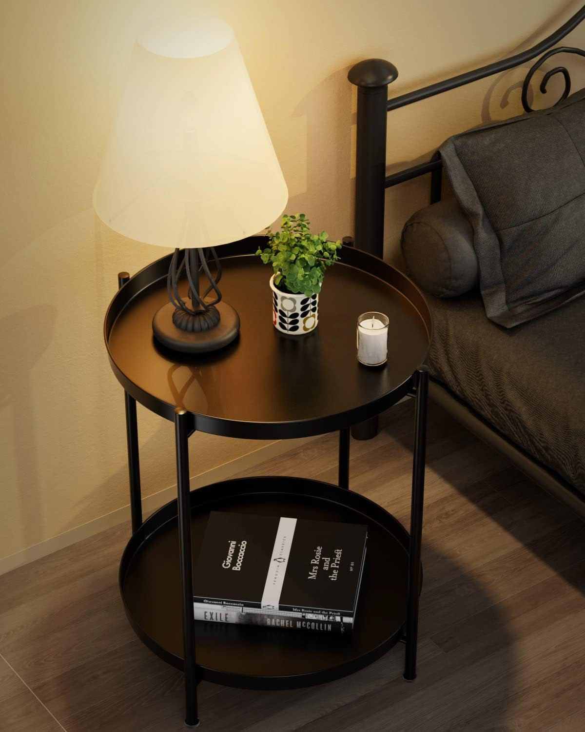 Black round side table beside a bed holding a lit lamp, small potted plant and candle, books on lower shelf.