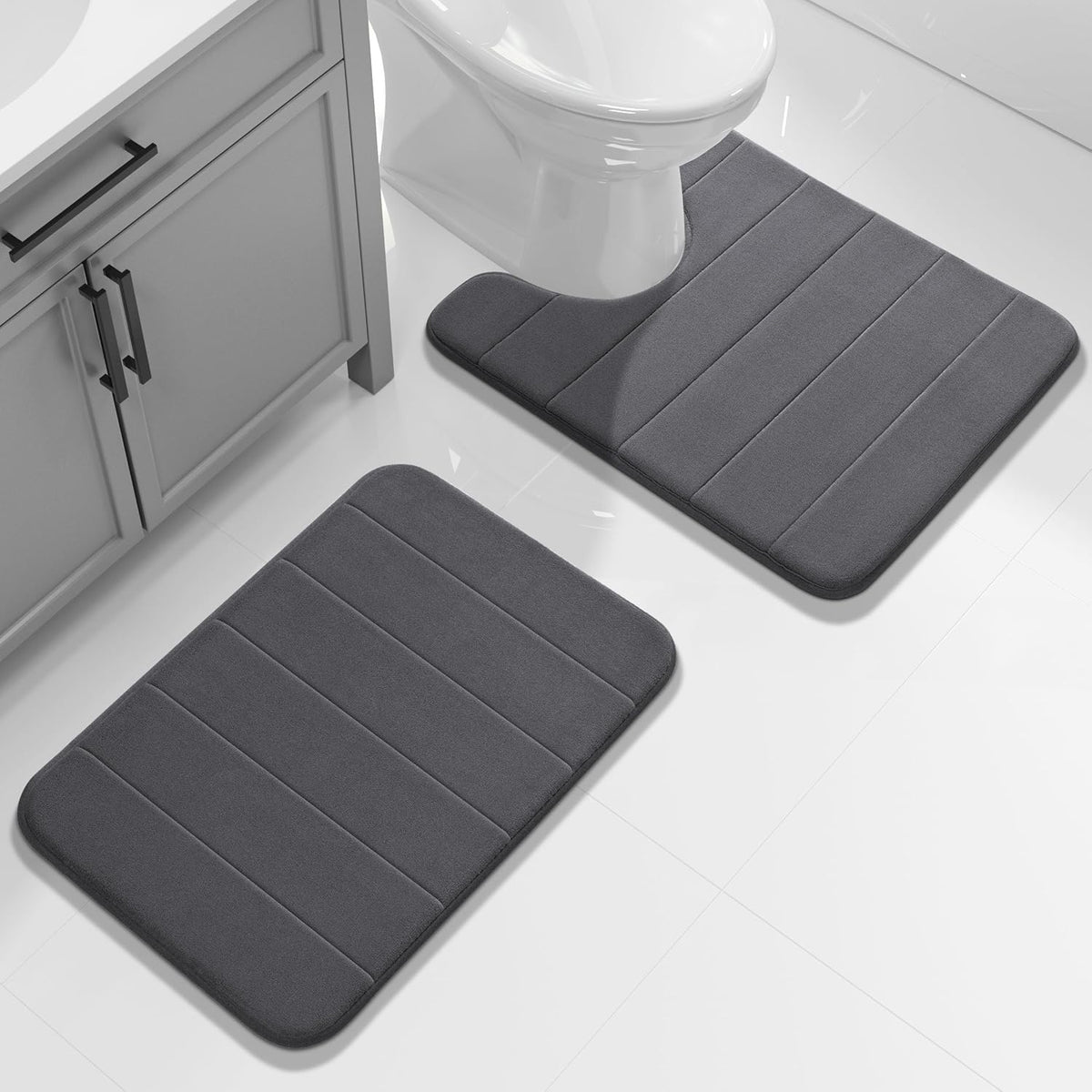 Two dark gray memory-foam bath mats on glossy white tile, one U-shaped around a toilet and one rectangular by a vanity