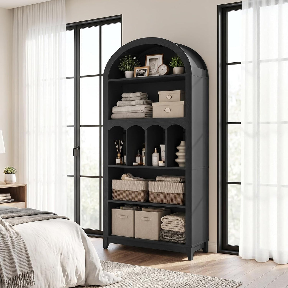 Side-front bedroom view of black arched bookcase beside a bed and tall windows, lower shelves with baskets visible.