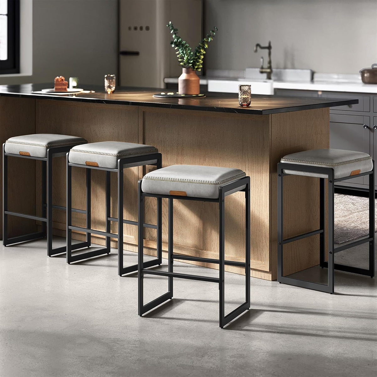 Four light-gray cushioned counter stools with black frames beside a kitchen island topped in dark stone.