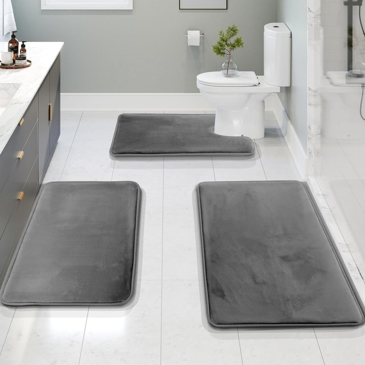 Three charcoal-gray bath rugs arranged on a white tile bathroom floor with a toilet and vanity.
