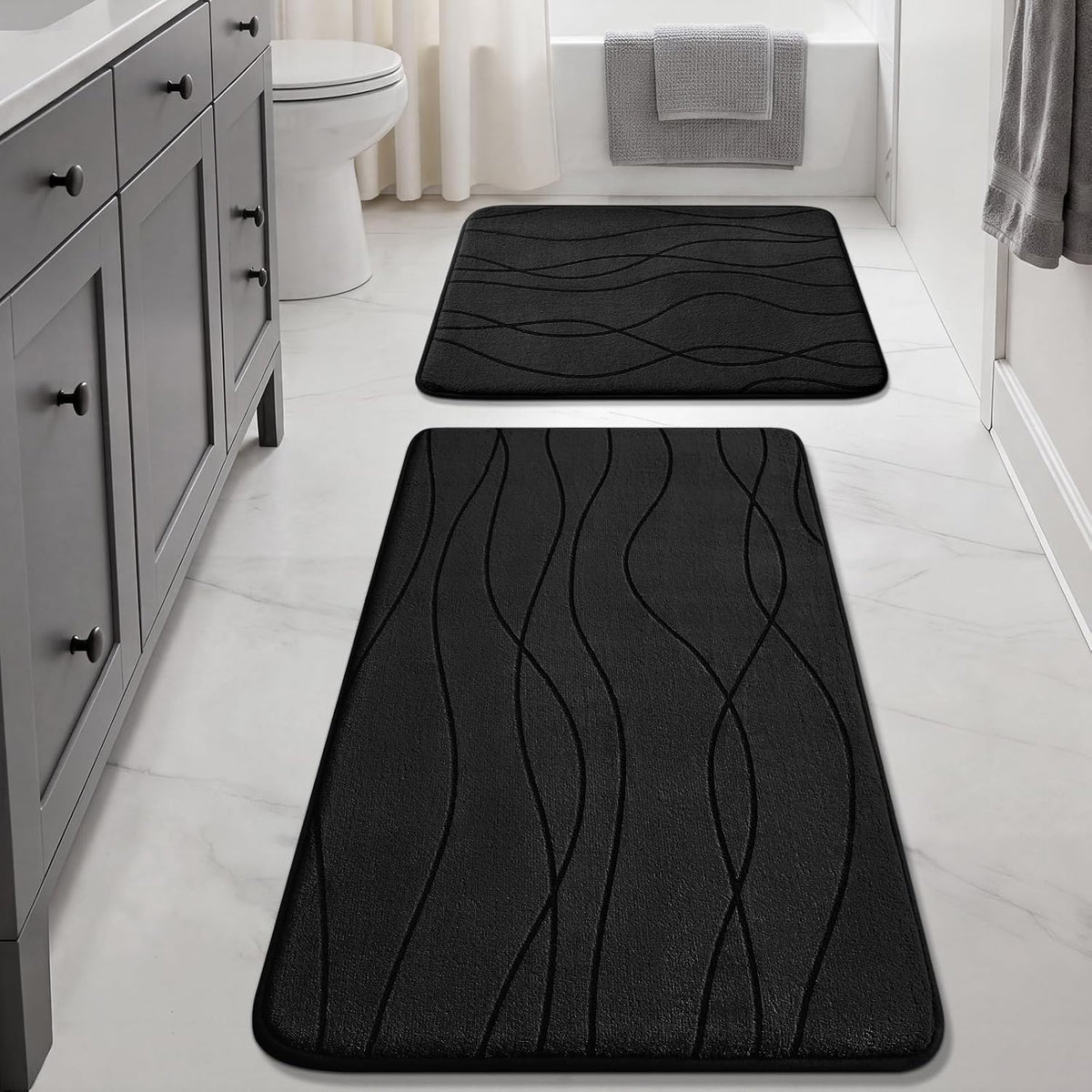 Two black bath mats with raised wavy lines placed on a white tile bathroom floor in front of a tub and vanity.