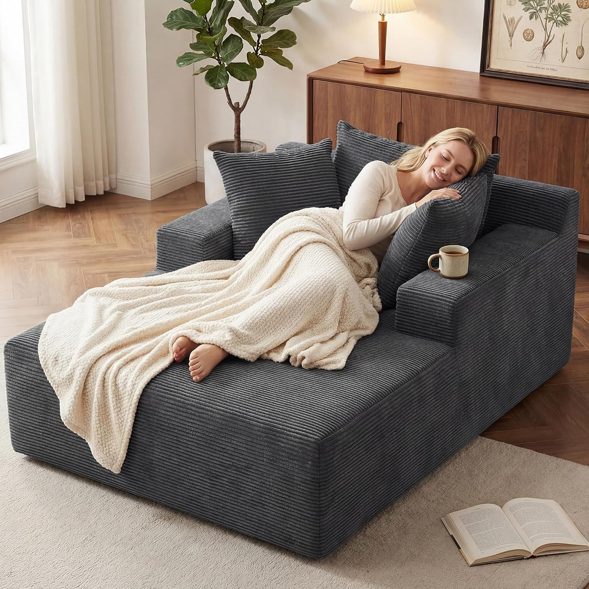 Three-quarter view of a dark gray oversized chaise with a woman hugging a pillow, mug on the arm, and an open book on the