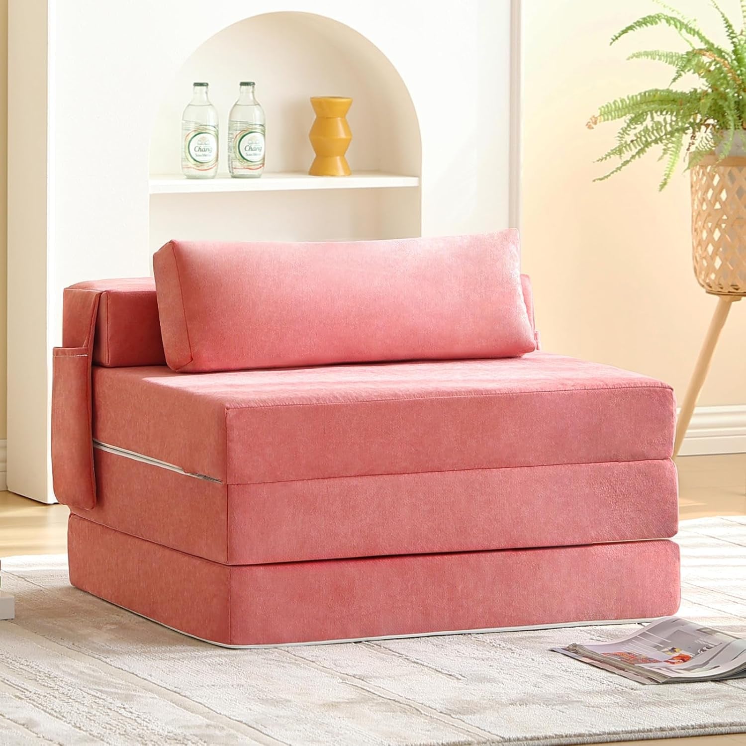 Coral pink foldable sofa bed chair displayed in a softly lit room with decorative shelf behind.