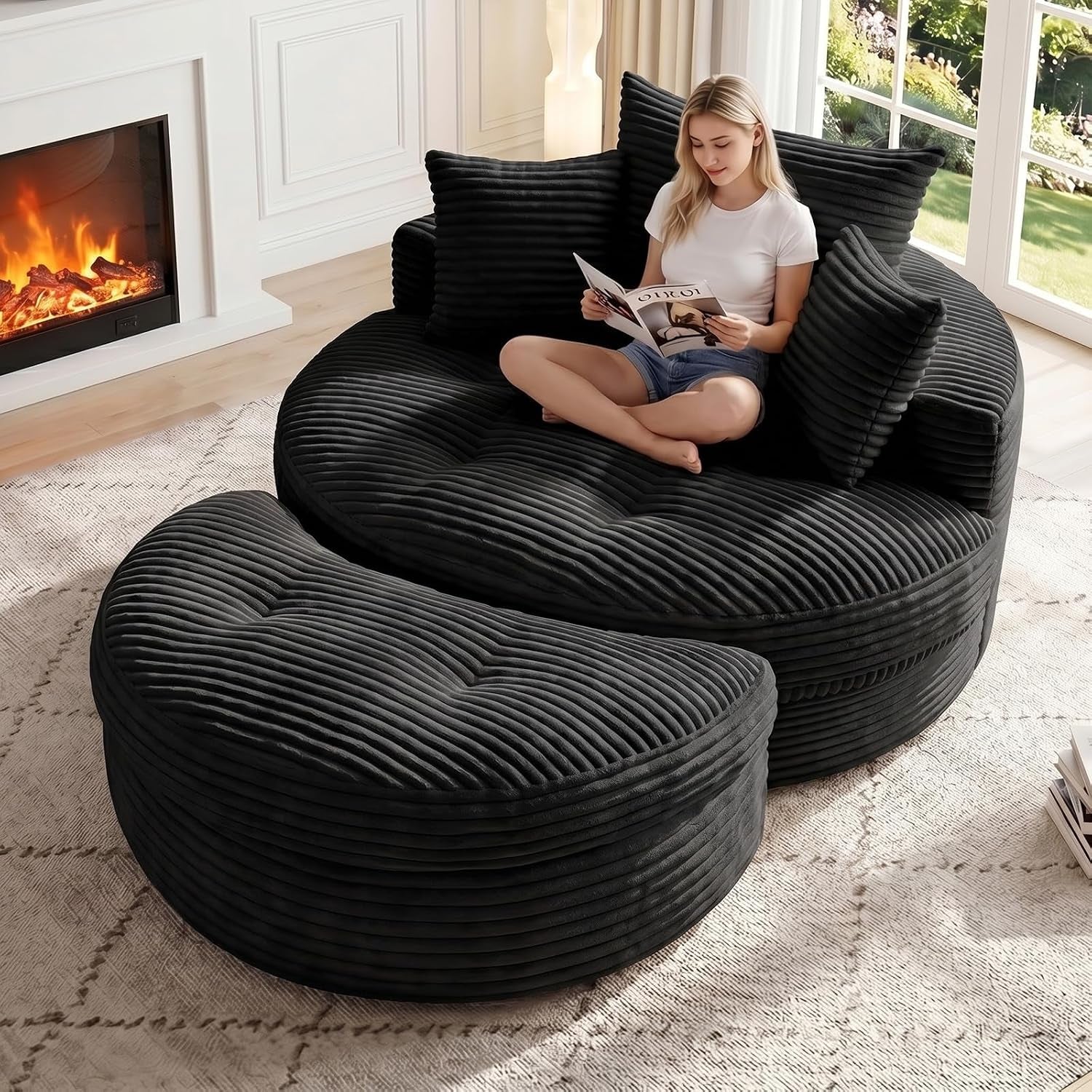 Angled view of the black round chaise with matching curved ottoman on a rug beside a fireplace; person seated reading.