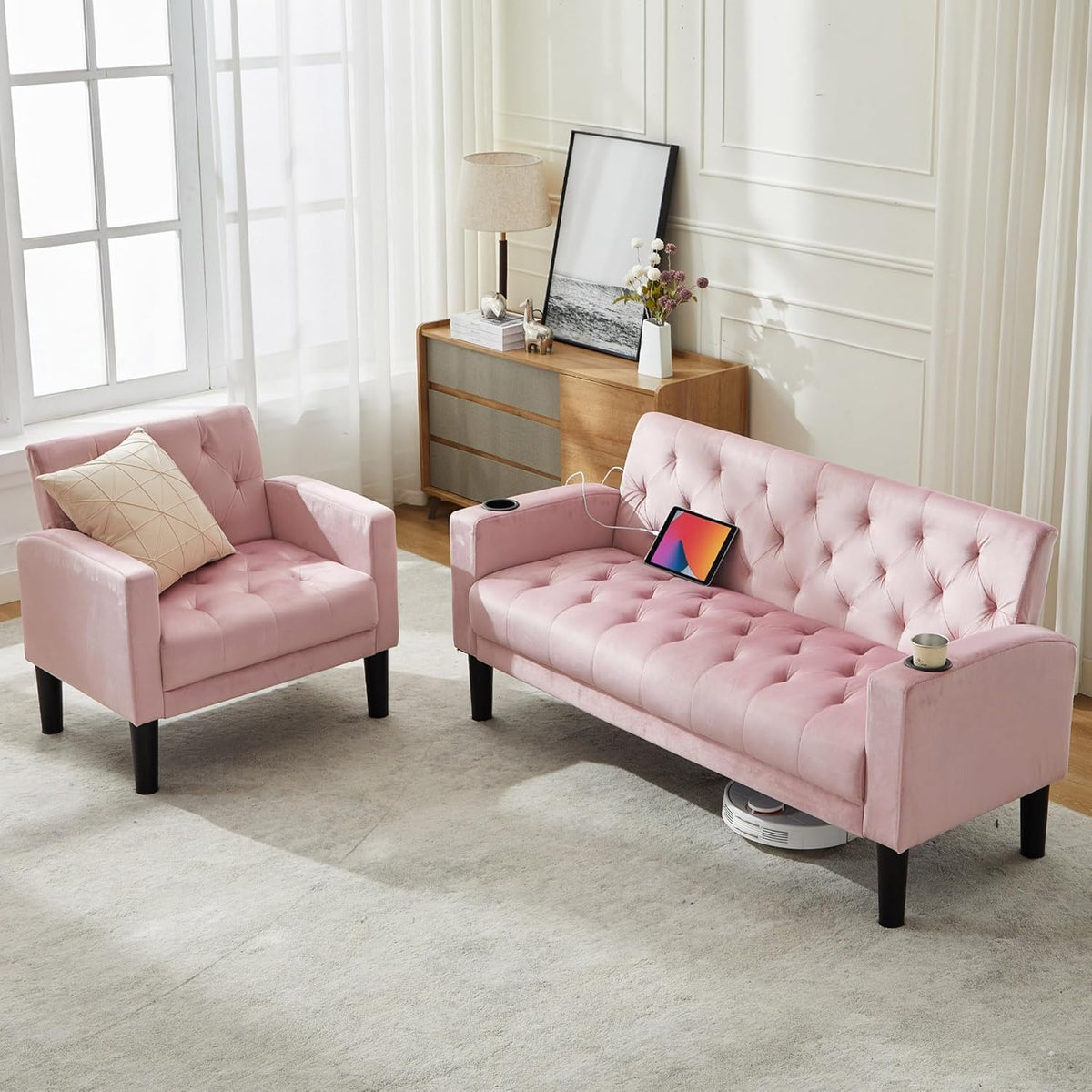 Angled view of a pink tufted loveseat and armchair beside a wooden dresser with lamp and framed art.