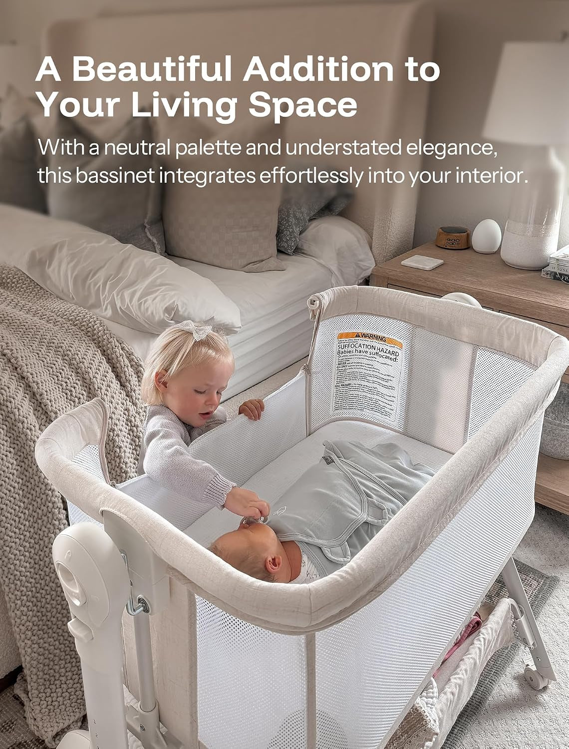 Beige bassinet next to a bed with a toddler reaching in to touch a newborn inside.