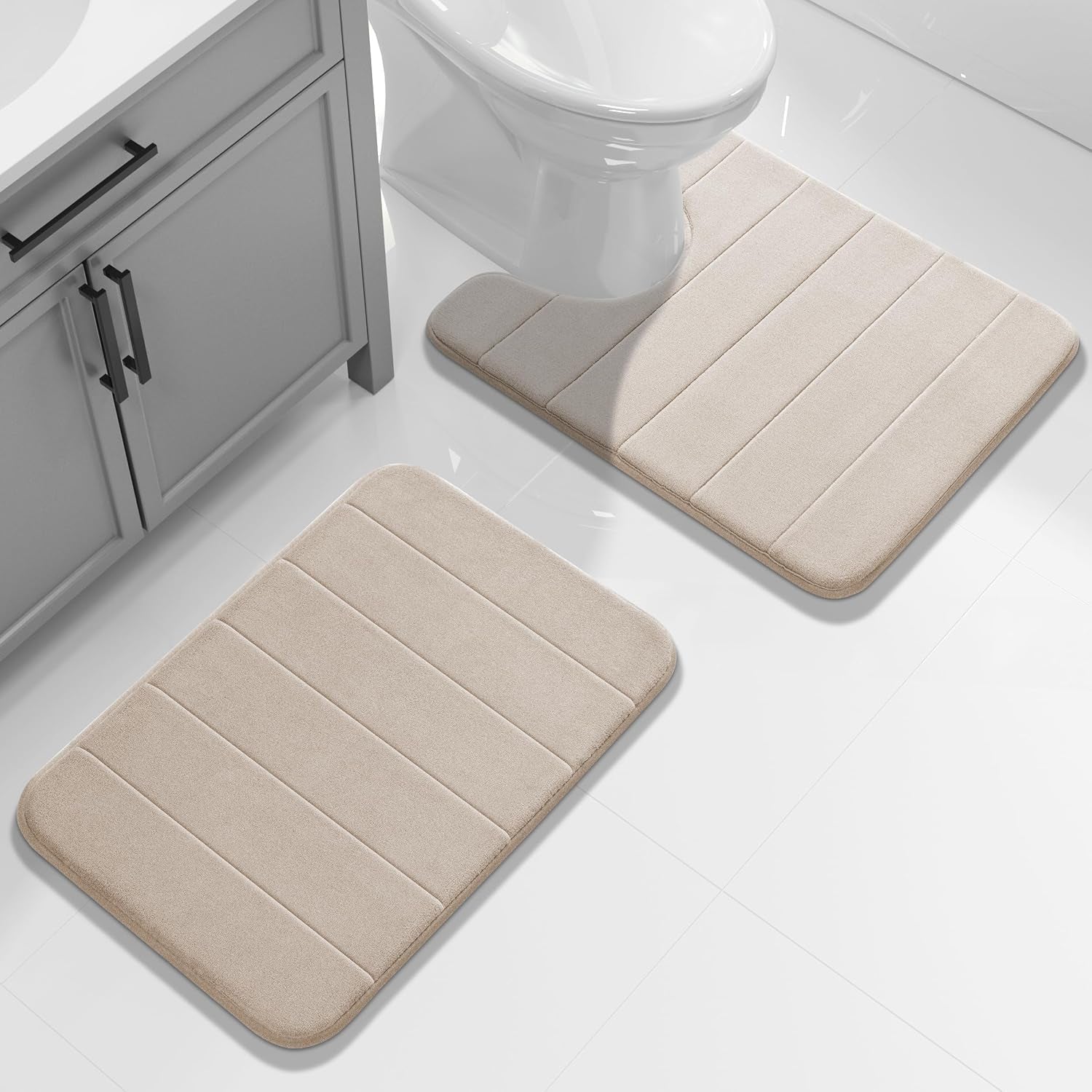 Two beige memory-foam bathroom mats on white tile placed near a vanity and toilet