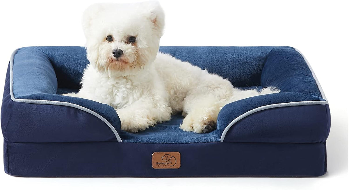 Navy blue bolster bed with light gray piping and a small white fluffy dog sitting upright on the sleeping surface.