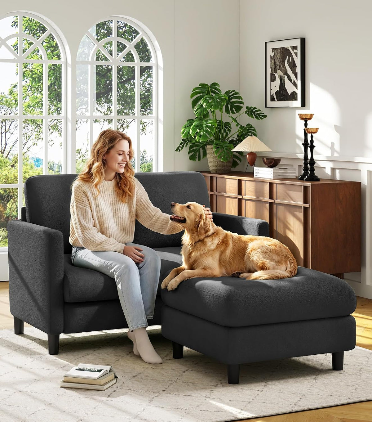 Woman petting a golden retriever on a dark gray two-piece loveseat and ottoman in a sunlit living room.