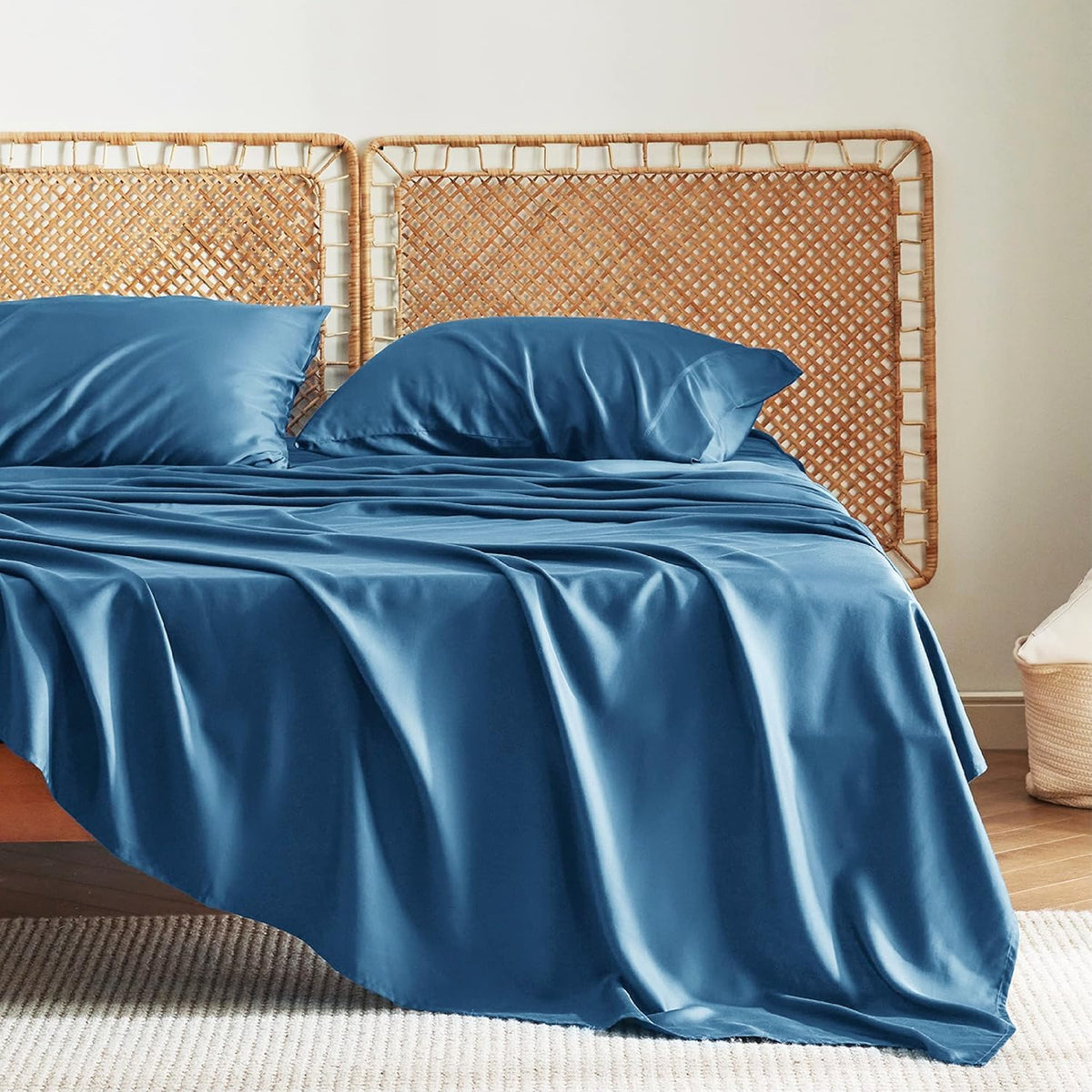 Three-quarter view of a bed with glossy cobalt-blue sheets and two pillows against a woven cane headboard.