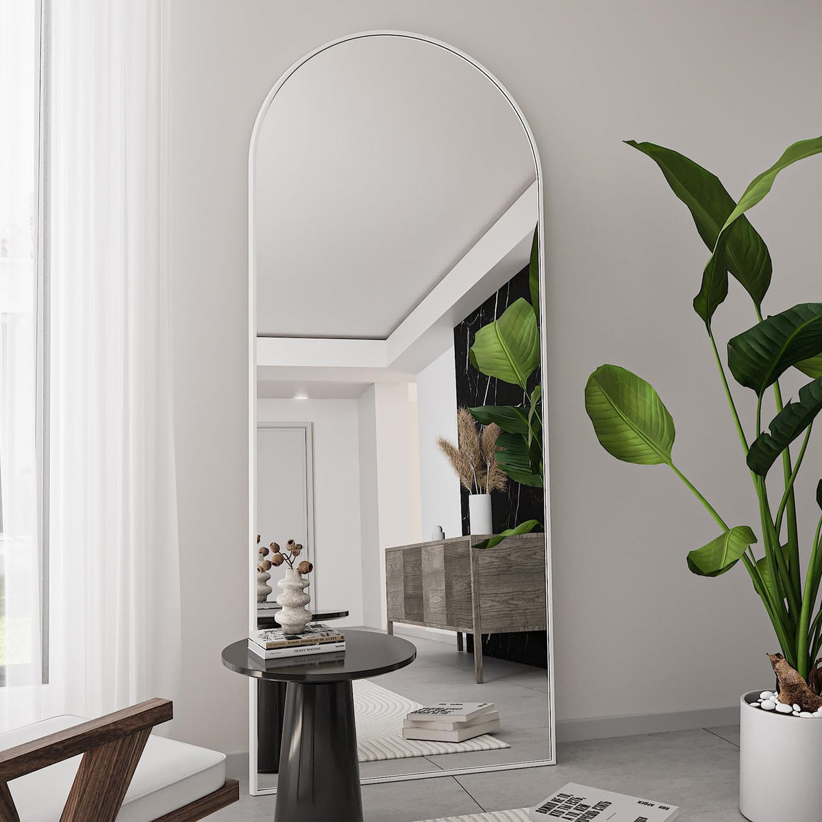 Arched full-length mirror with a slim white frame placed beside a round table and plant.