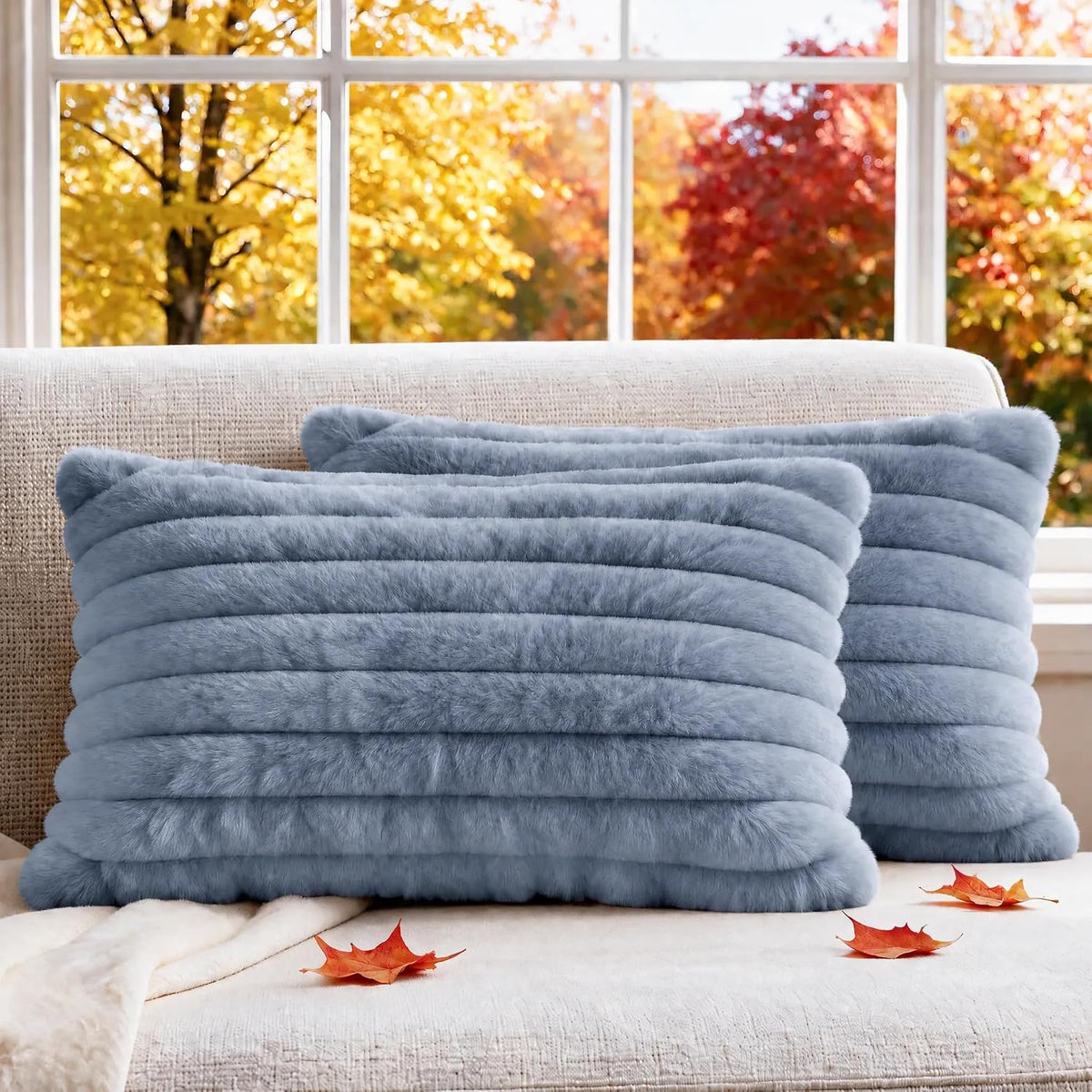 Pair of soft ribbed faux-fur pillows in pale blue placed on a couch with autumn foliage visible through the window.