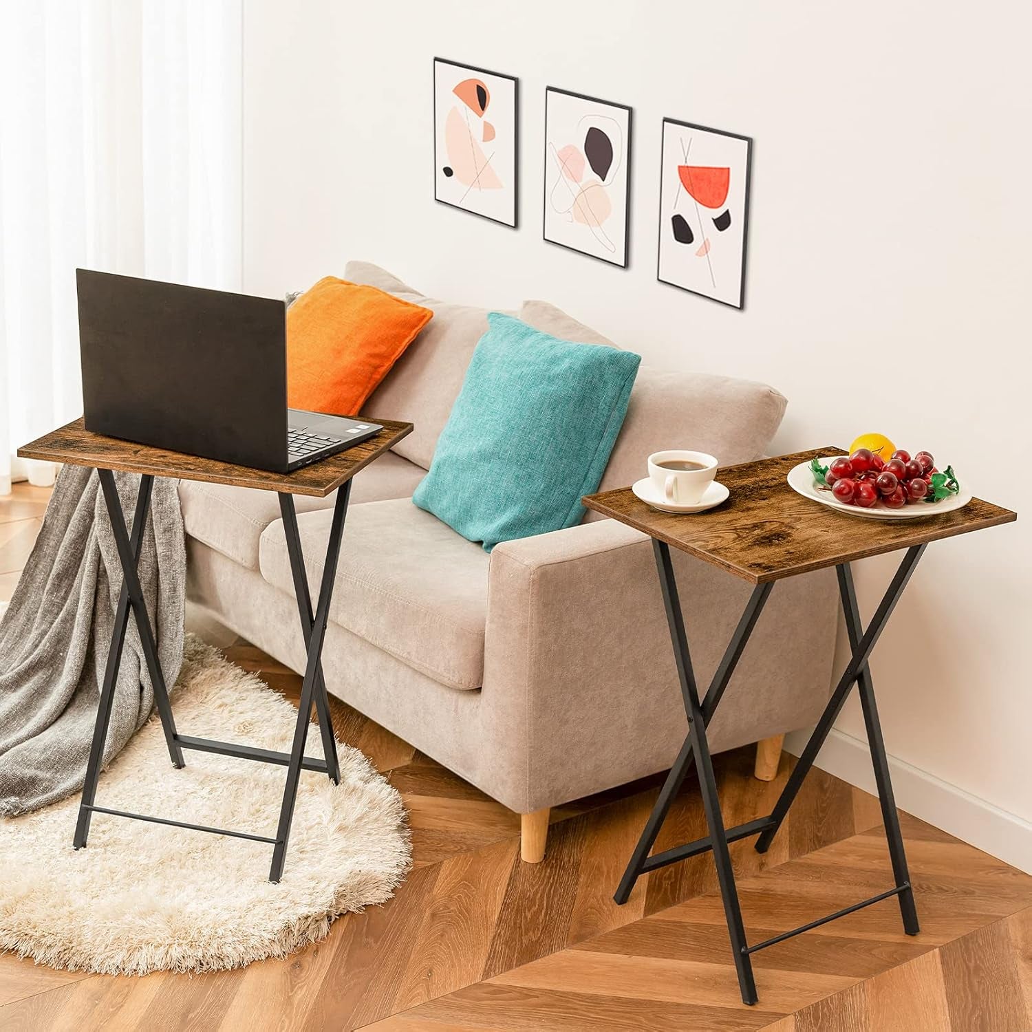 Two folding tray tables next to a couch; one holds a laptop, the other a cup and plate of fruit.