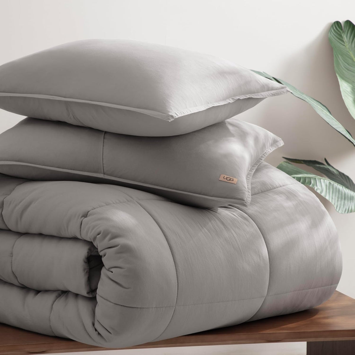 Rolled comforter with two matching shams stacked on a wooden bench next to a plant.