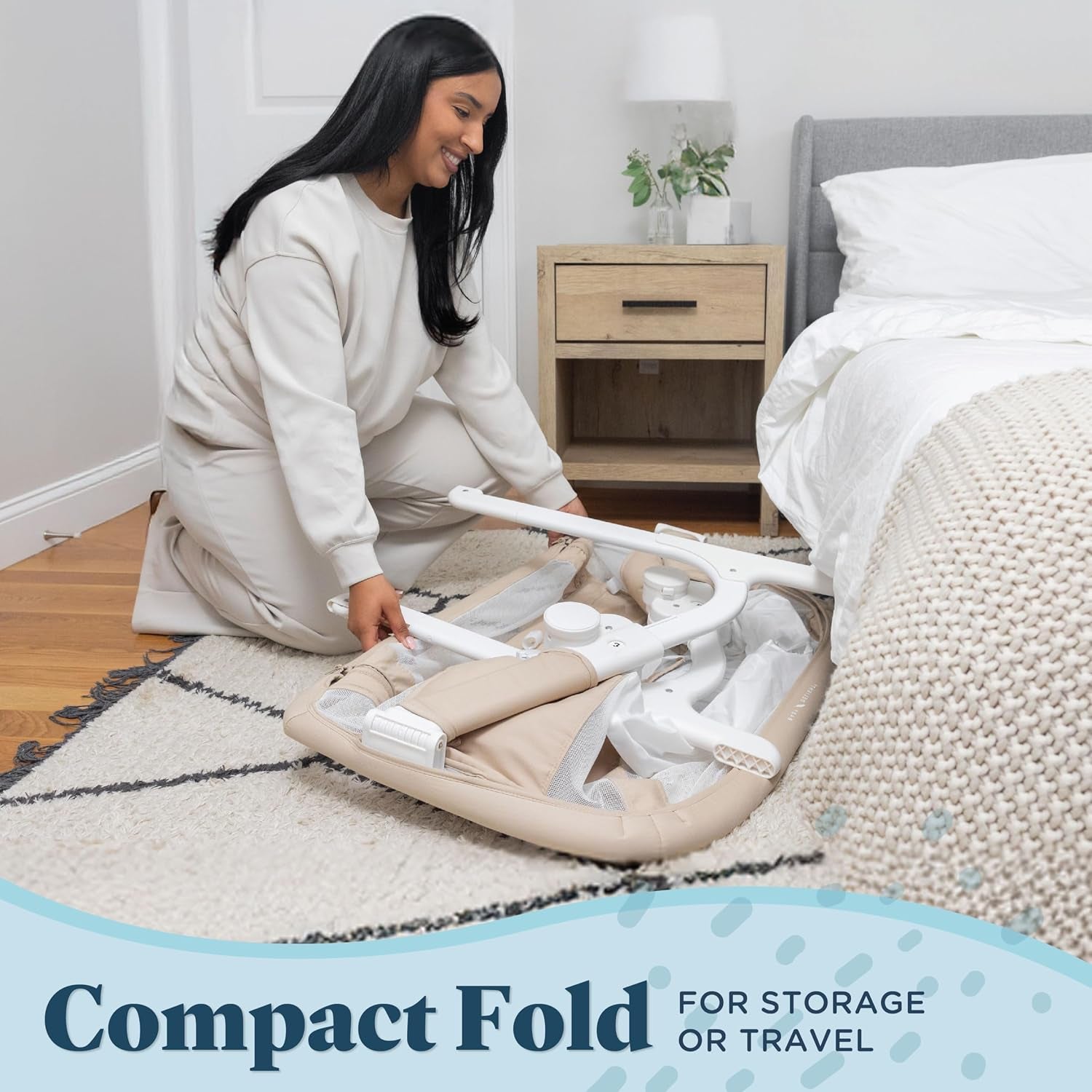 Side view of a woman folding the beige bassinet flat on a bedroom rug beside a bed.