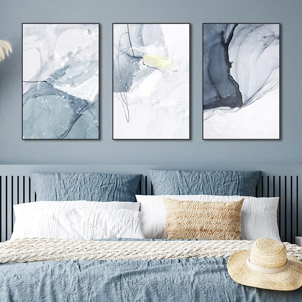 Three framed blue-gray abstract canvases arranged above a bed with blue pillows in a straight-on bedroom view