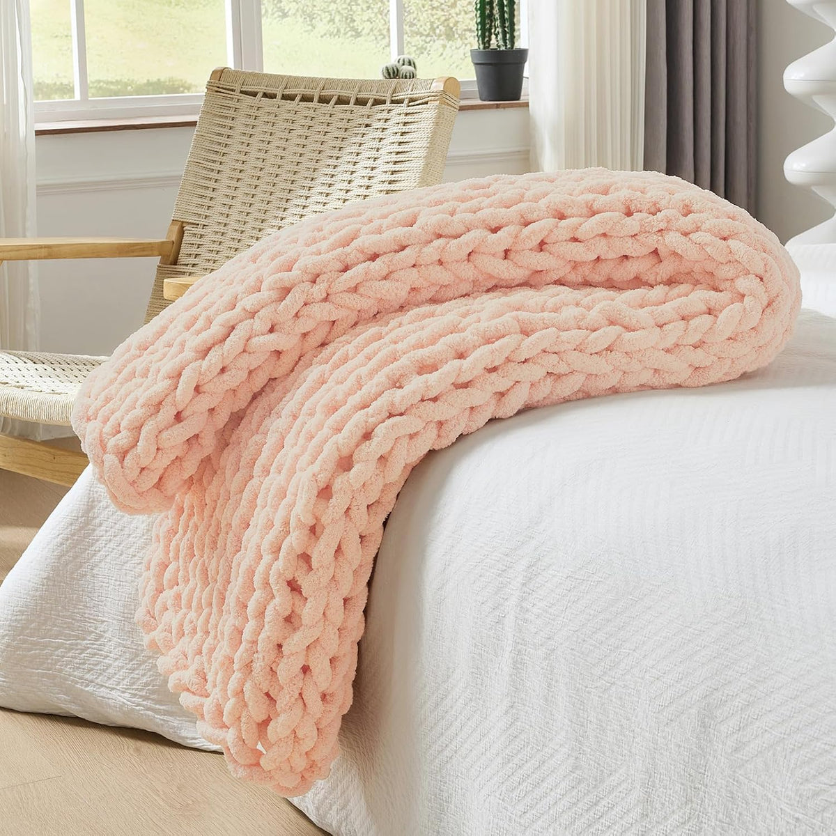 Blush-pink chunky knit throw casually folded and draped over the bed edge beside a woven chair.