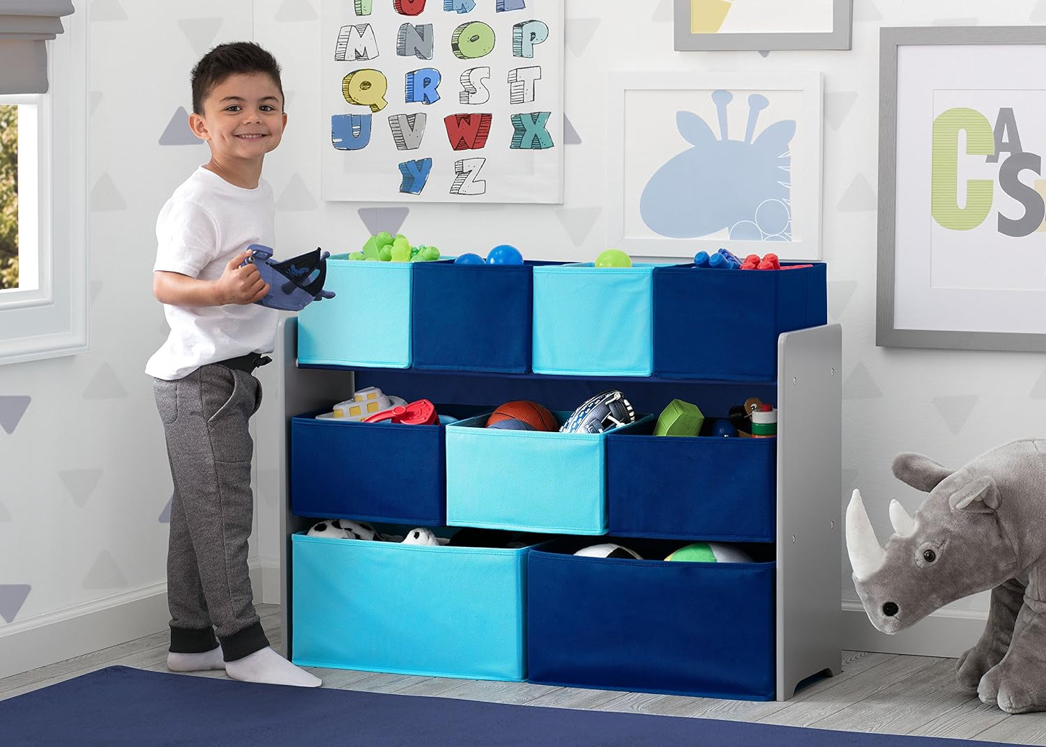 Main room shot - three-tier gray shelf with nine navy and aqua fabric bins filled with toys, child standing beside it.