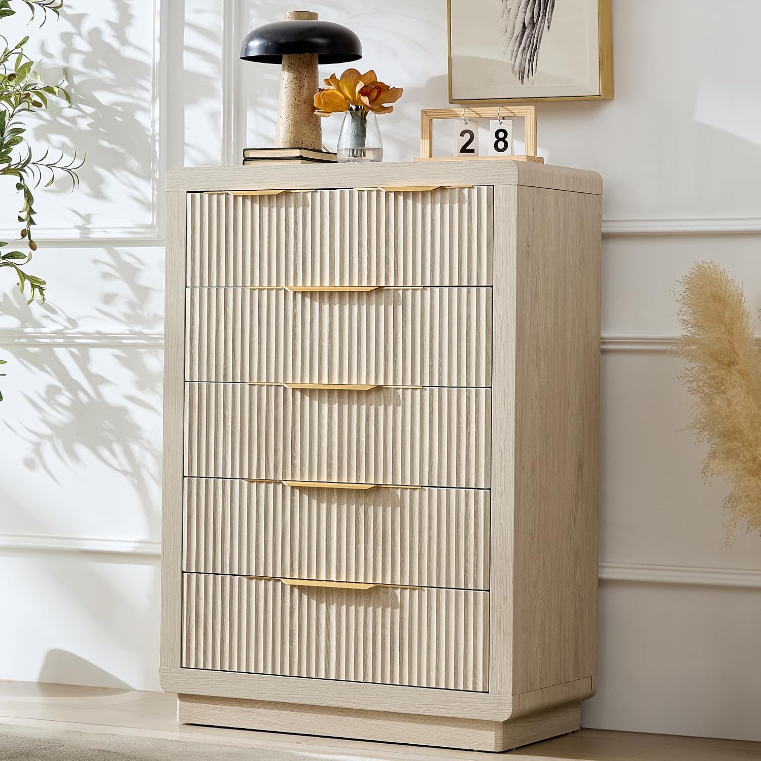 Tall ribbed-front chest in a light wood finish against a white wall, lamp and small decor on top.