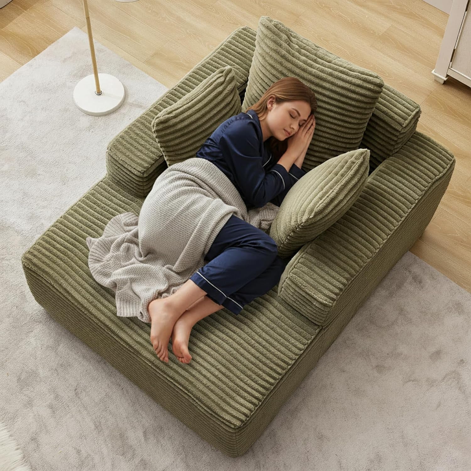 Overhead view of an olive-green corduroy chaise with three pillows and a woman curled up under a blanket.