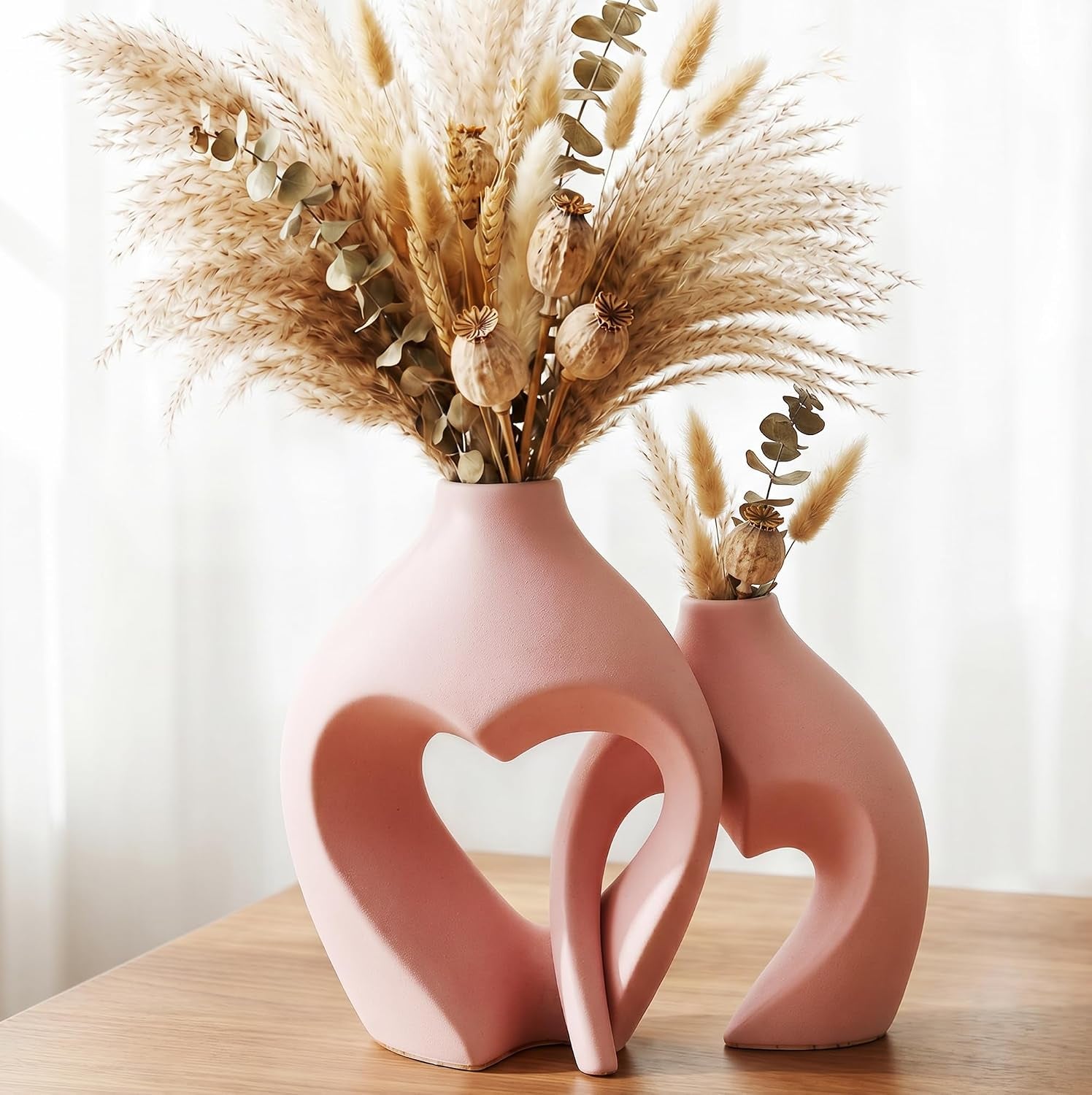 Pair of blush-pink heart vases on a wooden surface styled with fluffy dried pampas.