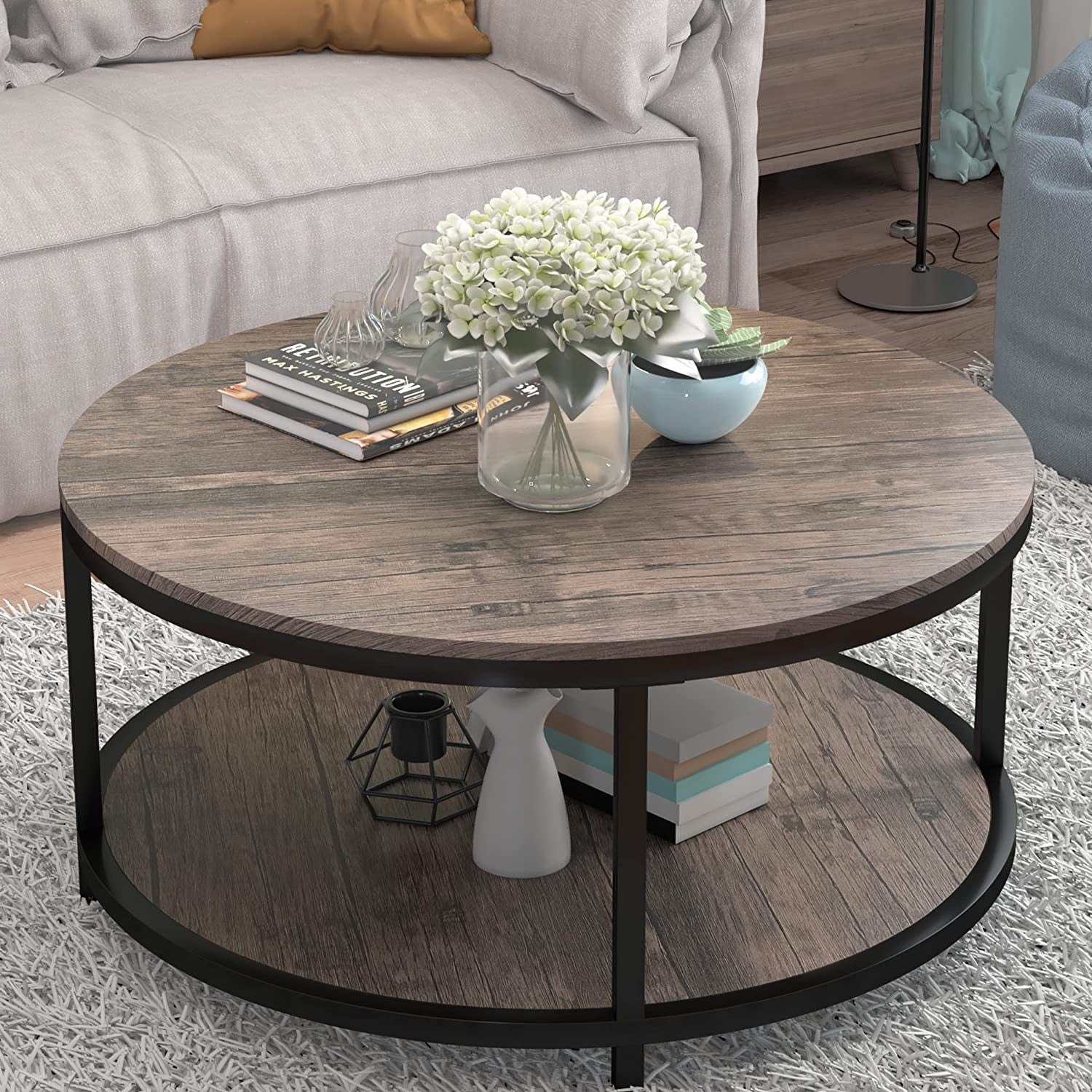 Round two-tier table with textured wood finish, vase of flowers on top and books on the lower shelf in a living room setting.