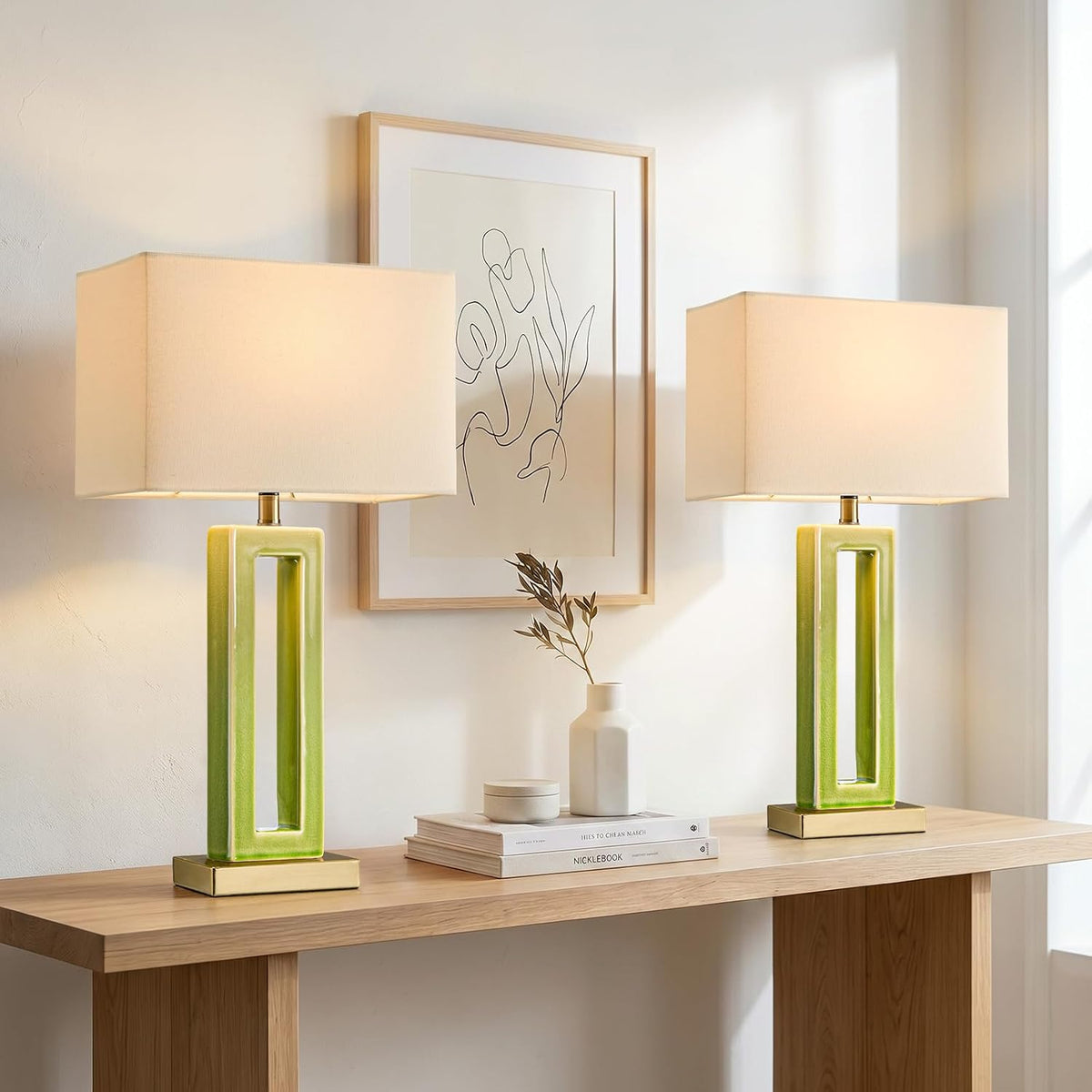 Two green crackle-glaze ceramic rectangular table lamps with beige shades on a light wood console under framed artwork