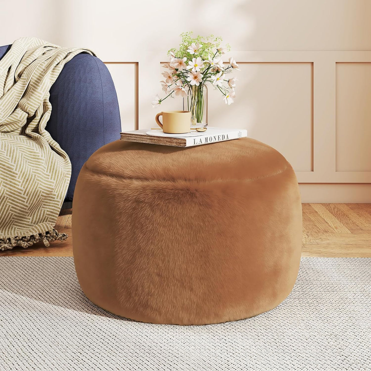 Orange-brown faux-fur round pouf ottoman with book and vase on top beside a sofa
