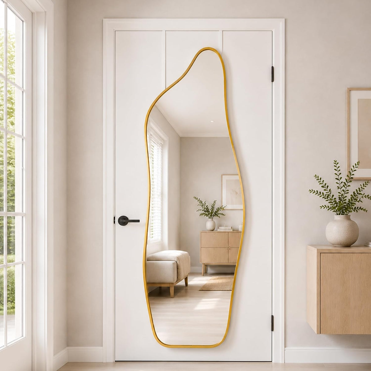 Full-length irregular arched mirror with a gold frame hanging on a white door reflecting a light living space.