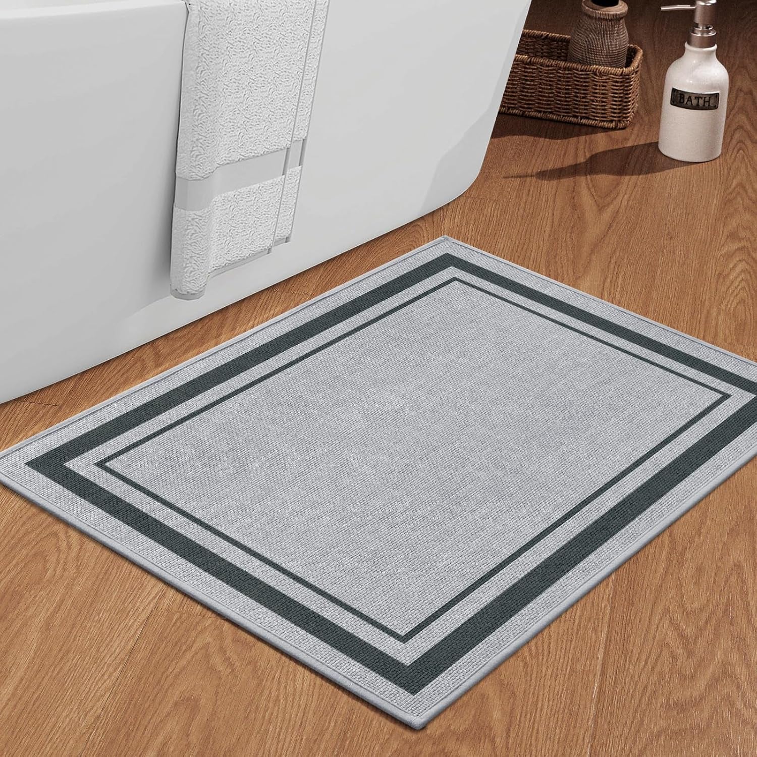 Light gray rectangular bath mat with double dark border on wood floor beside a tub