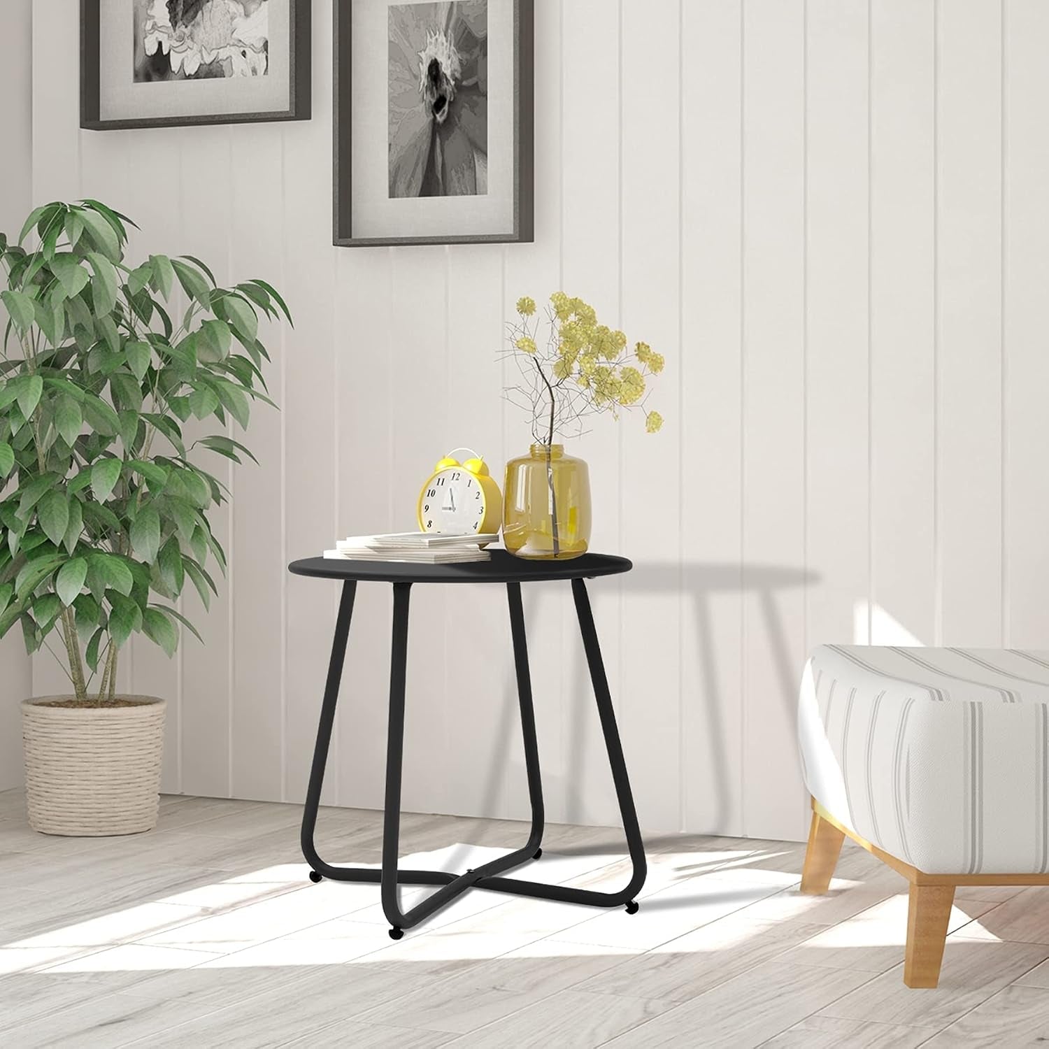 Black round metal side table styled beside a plant and ottoman with vase and alarm clock on top