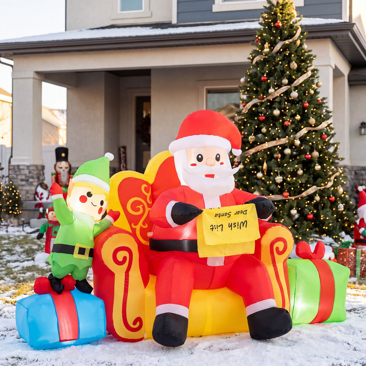 Inflatable Santa and an elf sit among gift boxes in the snow beside a lit Christmas tree.