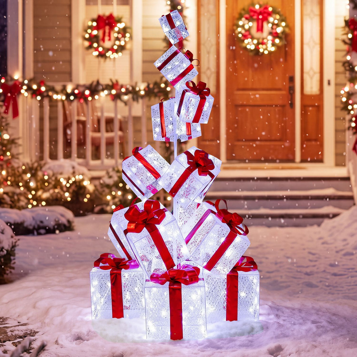 A lit stack of white gift boxes tied with red bows sits in the snow outside a decorated home.