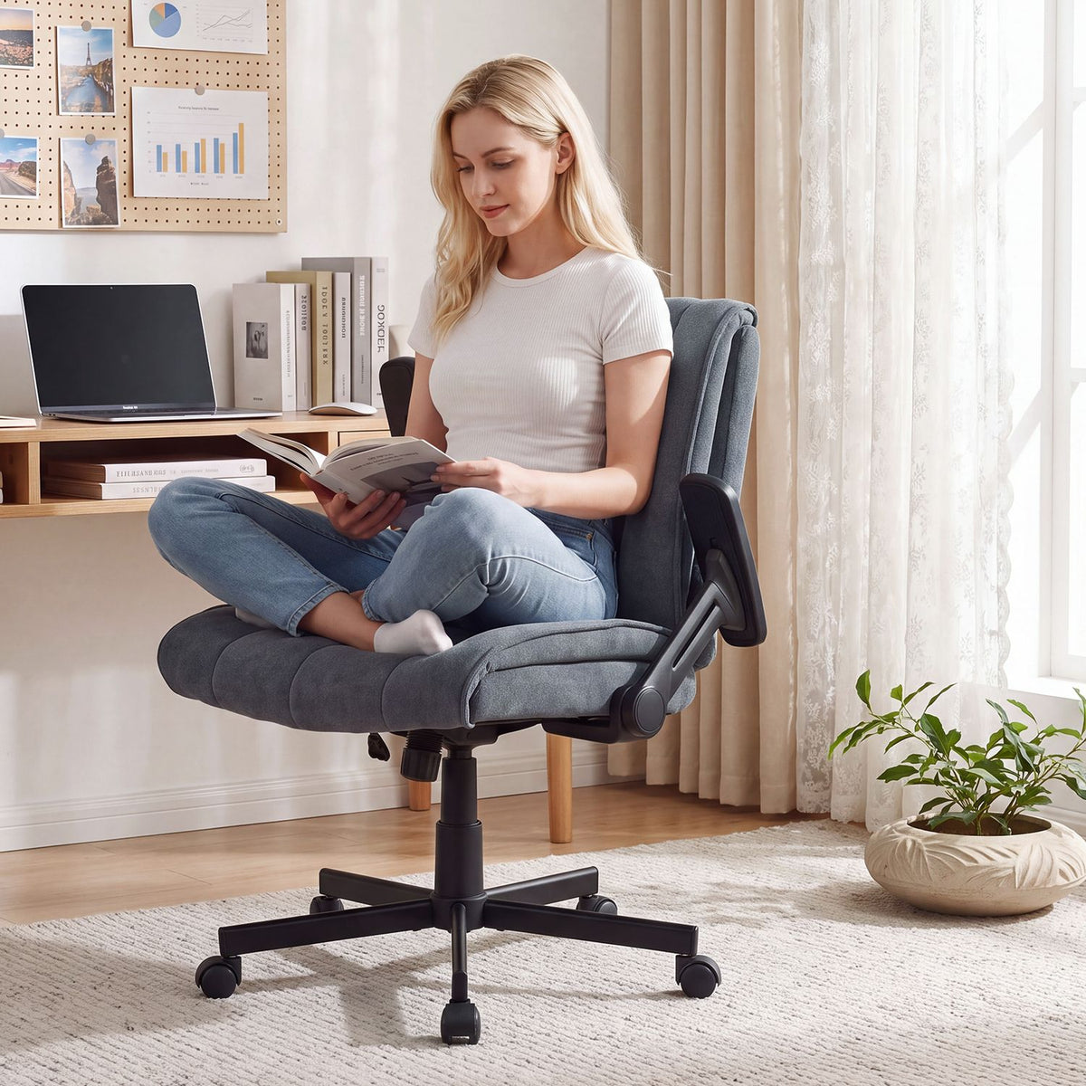 A woman sits cross-legged reading in a gray office chair beside a bright home-office desk.