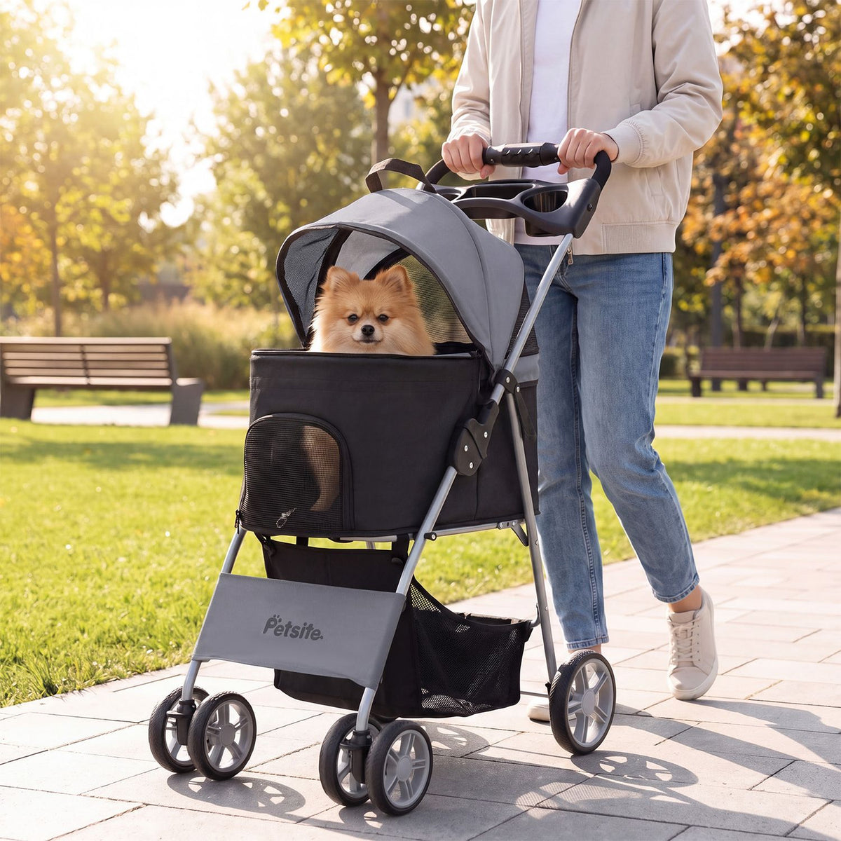 A person pushes a gray-and-black pet stroller with a small dog seated inside along a sunny park path.