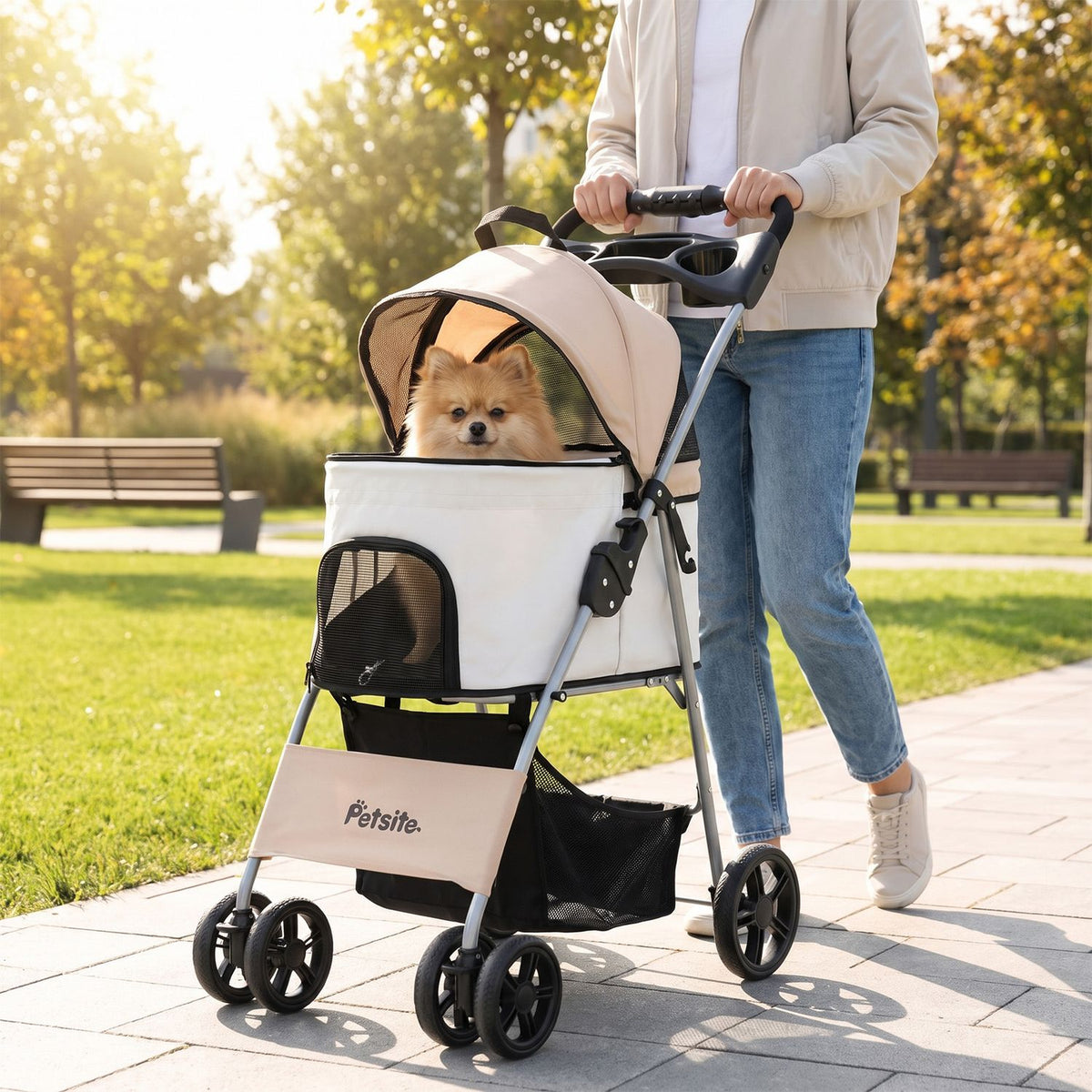 A person pushes a beige-and-white pet stroller with a small dog inside along a sunny park path.