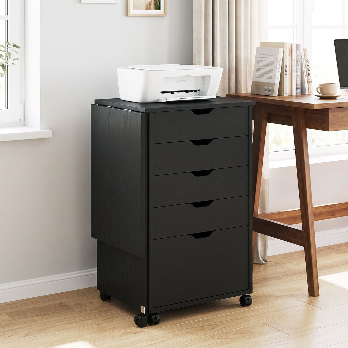 Black desk folded into a compact drawer cabinet with a printer on top, beside a wooden table.