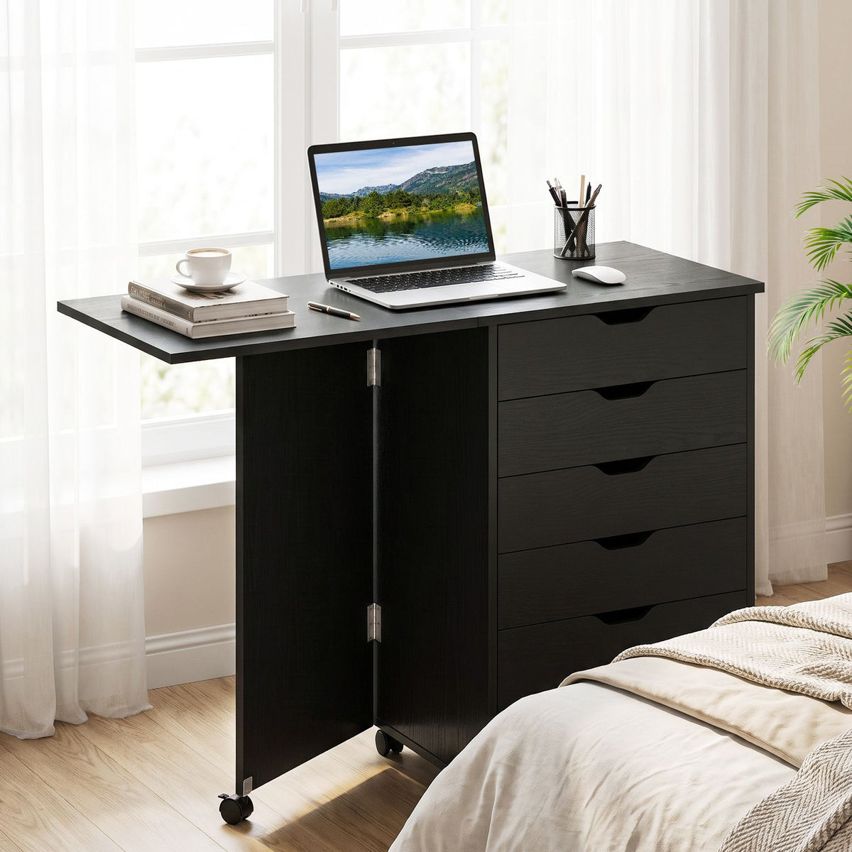 Open black folding desk with a laptop and books on top beside a bright bedroom window.