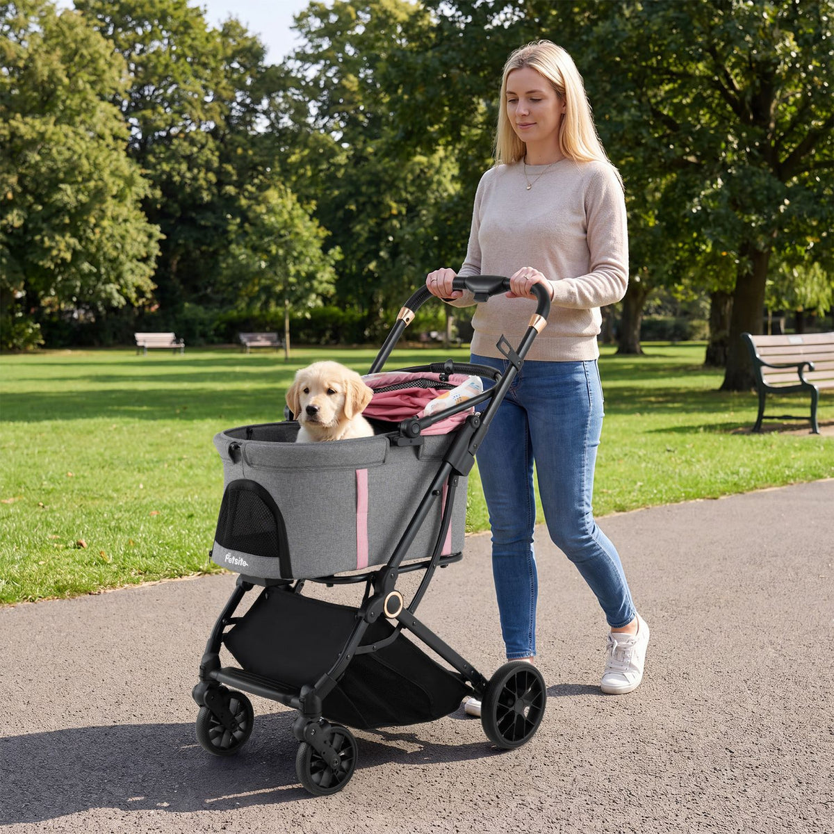A woman pushes a gray pet stroller with pink trim while a small dog sits inside in a sunny park.