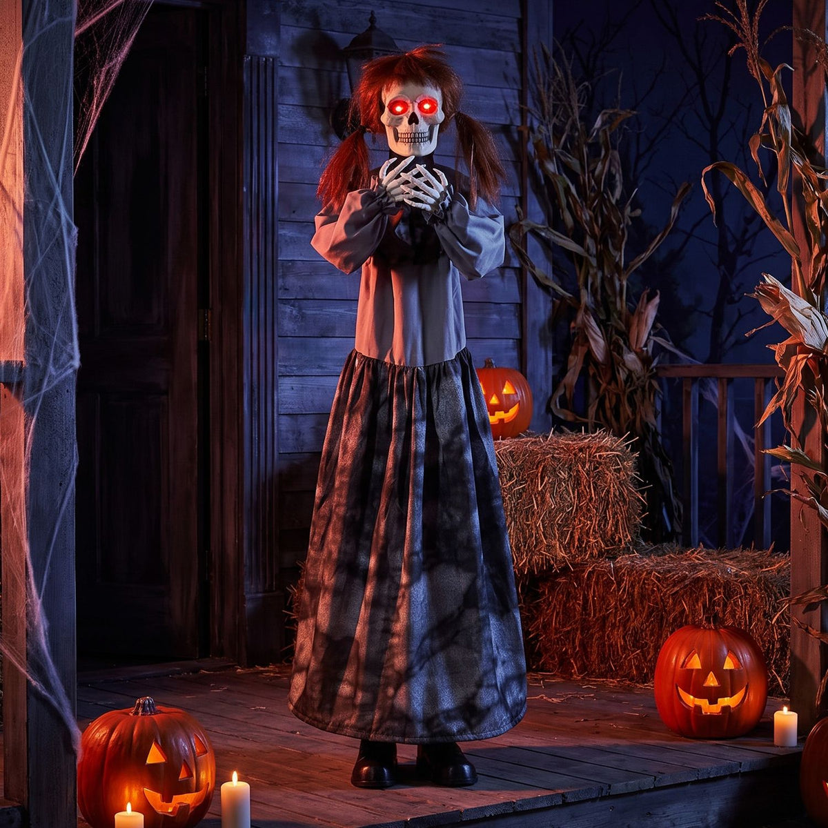 A red-eyed skeleton doll stands on a porch among glowing pumpkins, hay bales and dried cornstalks.