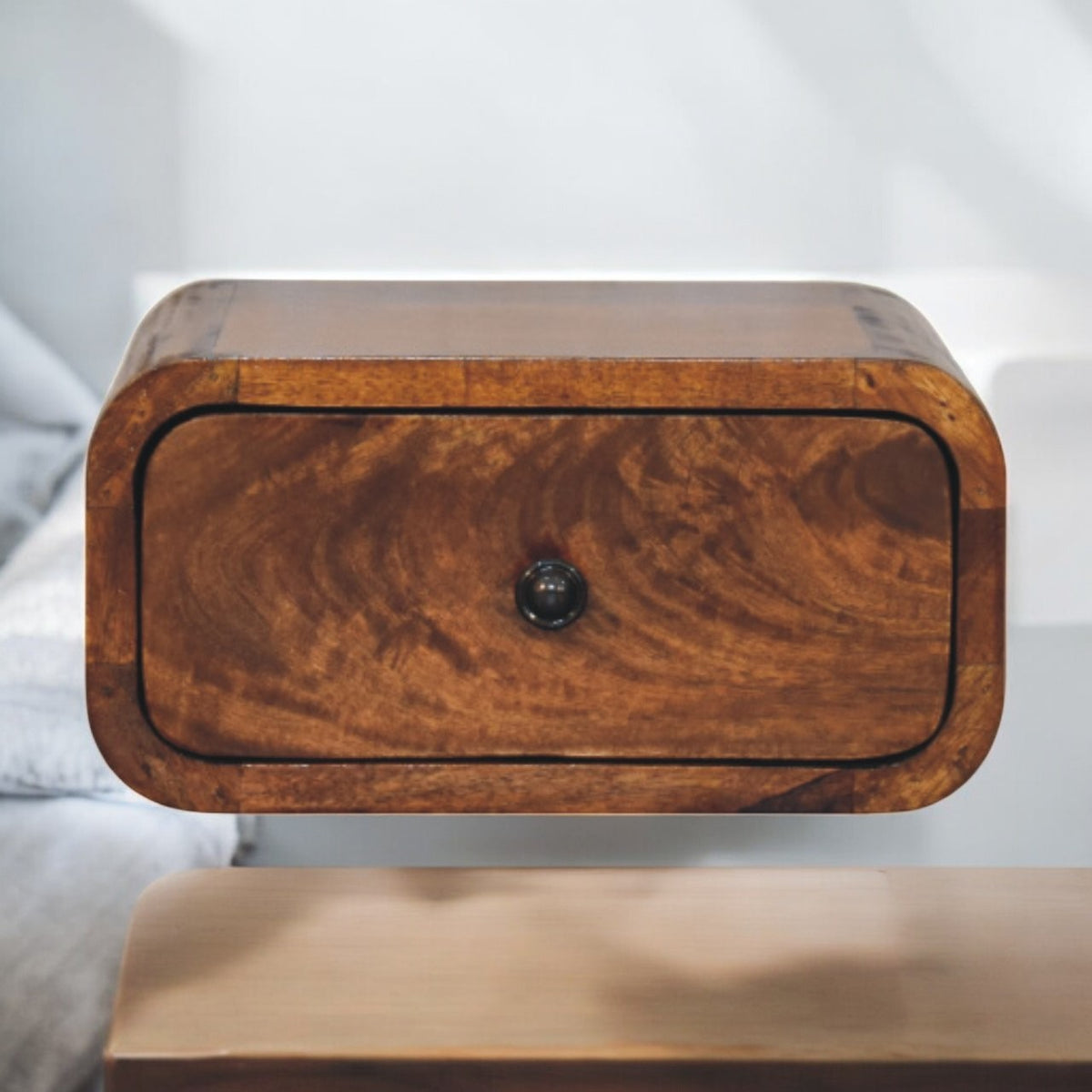 Wall-mounted rounded wooden nightstand with a single drawer and dark knob, shown beside a bed.