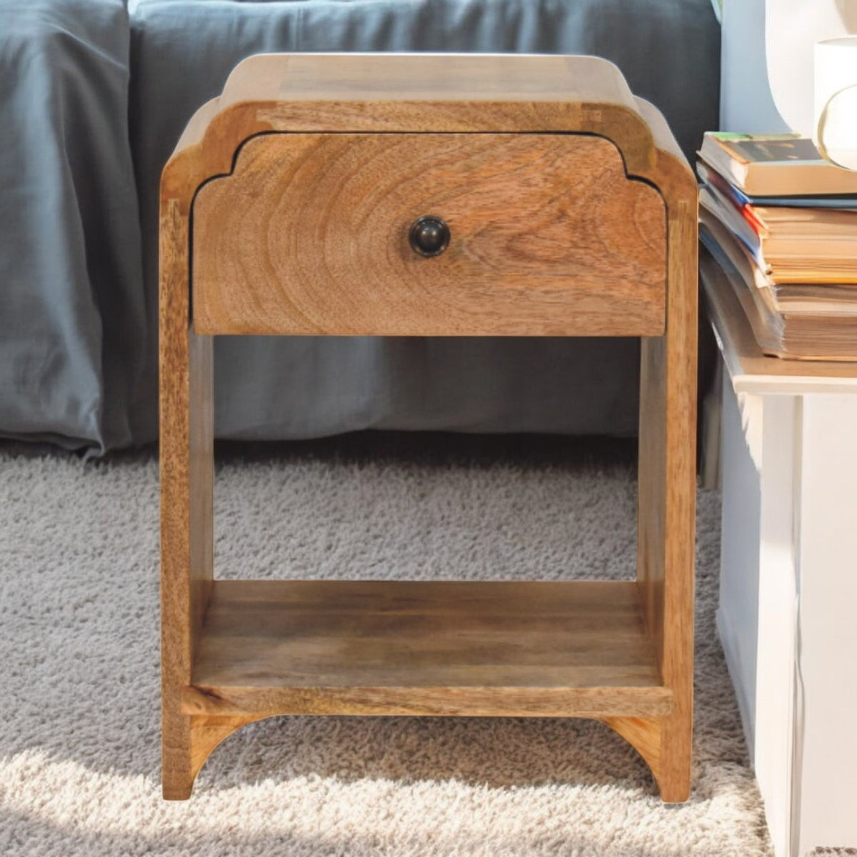 Light wood nightstand beside a bed, showing its drawer, dark knob, and open shelf.